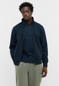 Giacca zip-up blu navy con collo alto, realizzata in materiale morbido. Abbinata a una maglia scura attillata e pantaloni verde chiaro, che mostrano uno stile casual.