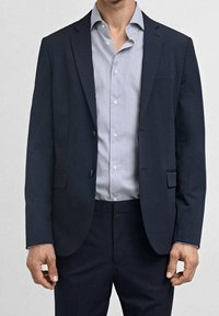 Laivaston sininen bleiseri, jossa on lovetut kaulukset, kaksi etutaskua ja yhden napin kiinnitys. Yllä vaaleansininen ja valkoinen raidallinen nappipaita.