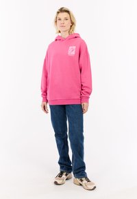 Hoodie en coton rose avec détail de poche avant, coupe décontractée et capuche avec cordon de serrage, associé à un jean bleu foncé et des chaussures de sport beiges.