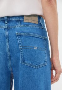 Blå denimjeans med hög midja, bakfickor och en brun läderlapp på midjebandet med präglad text.