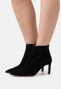 Botas de tobillo de ante negro con punta afilada y un diseño elegante. Cuentan con un tacón de aguja y un sutil acento dorado en el lateral.