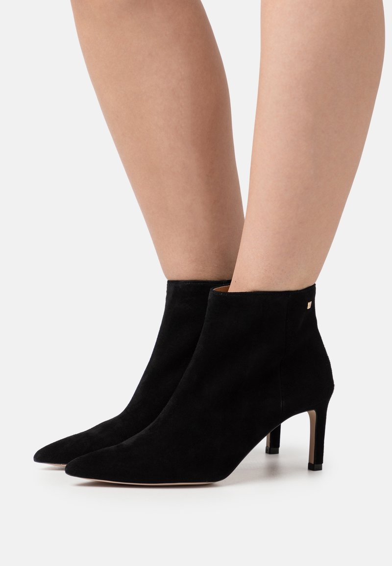 Botas de tobillo de ante negro con punta afilada y un diseño elegante. Cuentan con un tacón de aguja y un sutil acento dorado en el lateral.