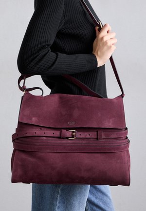 Una persona che indossa un maglione a coste nero e jeans blu tiene in mano una grande borsa a spalla in suede bordeaux con dettagli in metallo sulla parte anteriore.