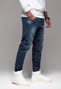 Jogger in denim blu con tasche alle ginocchia, vestibilità slim, polsini elasticizzati e dettagli in zip dorati; abbinati a sneakers alte bianche.