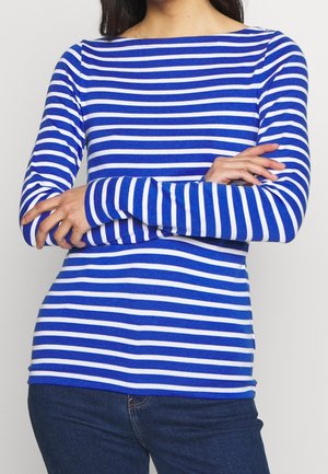 Persoon draagt een blauw-wit horizontaal gestreept shirt met lange mouwen en gekruiste armen, gecombineerd met donkerblauwe spijkerbroek.