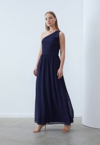 Tumši zila, vienpusēja maxi kleita ar savāktu jostas vietu un plūstošu svārku, izgatavota no viegla materiāla. Valkāta ar nude augstpapēžu sandālēm.