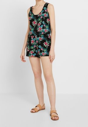 Kvinde iført en sort ærmeløs blomsterdragt med røde, blå og grønne blomster, kombineret med beige flade sandaler, stående mod en ensfarvet baggrund.