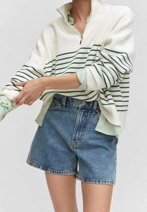 Person wearing a white sweater with green stripes, light green shirt underneath, and high-waisted blue denim shorts, adjusting one sleeve.