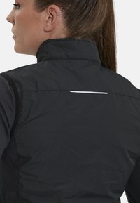Schwarze ärmellose Jacke mit hohem Kragen, strukturiertem Stoff und reflektierendem Streifen auf der Rückseite. Entwickelt für aktive Nutzung.