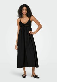 Zwarte maxi-jurk met dunne bandjes, gerimpelde lijfje en een zwierige rok. Gepaarde met zwarte slingback-schoenen met studs.