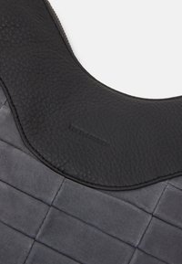 Svart läderaxelrem med en strukturerad yta och synliga sömmar, samt en slät, grå vadderad sektion. Logotypen är präglad.