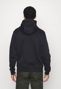 Svart hoodie med en stor, justerbar huva, tillverkad av mjukt tyg. Har långa ärmar och ribbade muddar, i kombination med olivgröna byxor.