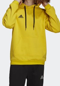 Felpa gialla con cappuccio e cordini neri, tasca a marsupio e logo adidas nero sulla parte superiore sinistra del petto. Tessuto morbido, vestibilità comoda.