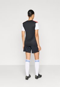 Μαύρη αθλητική φανέλα με λευκά μανίκια και κόκκινες λεπτομέρειες, συνοδευόμενη από σκούρο σορτς, λευκές κάλτσες μέχρι τα γόνατα με μπλε ρίγες, και μαύρα ποδοσφαιρικά παπούτσια.