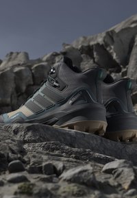 Czarno-turkusowe buty trekkingowe mają siateczkową cholewkę, grubą gumową podeszwę oraz trzy ukośne paski po bocznej stronie, umiejscowione na skalistym terenie.