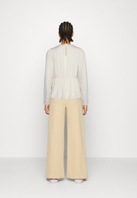 Blusa cremosa a maniche lunghe con vita elastica e dettaglio a chiave nella parte posteriore, abbinata a pantaloni a gamba larga beige chiaro e scarpe lavanda.