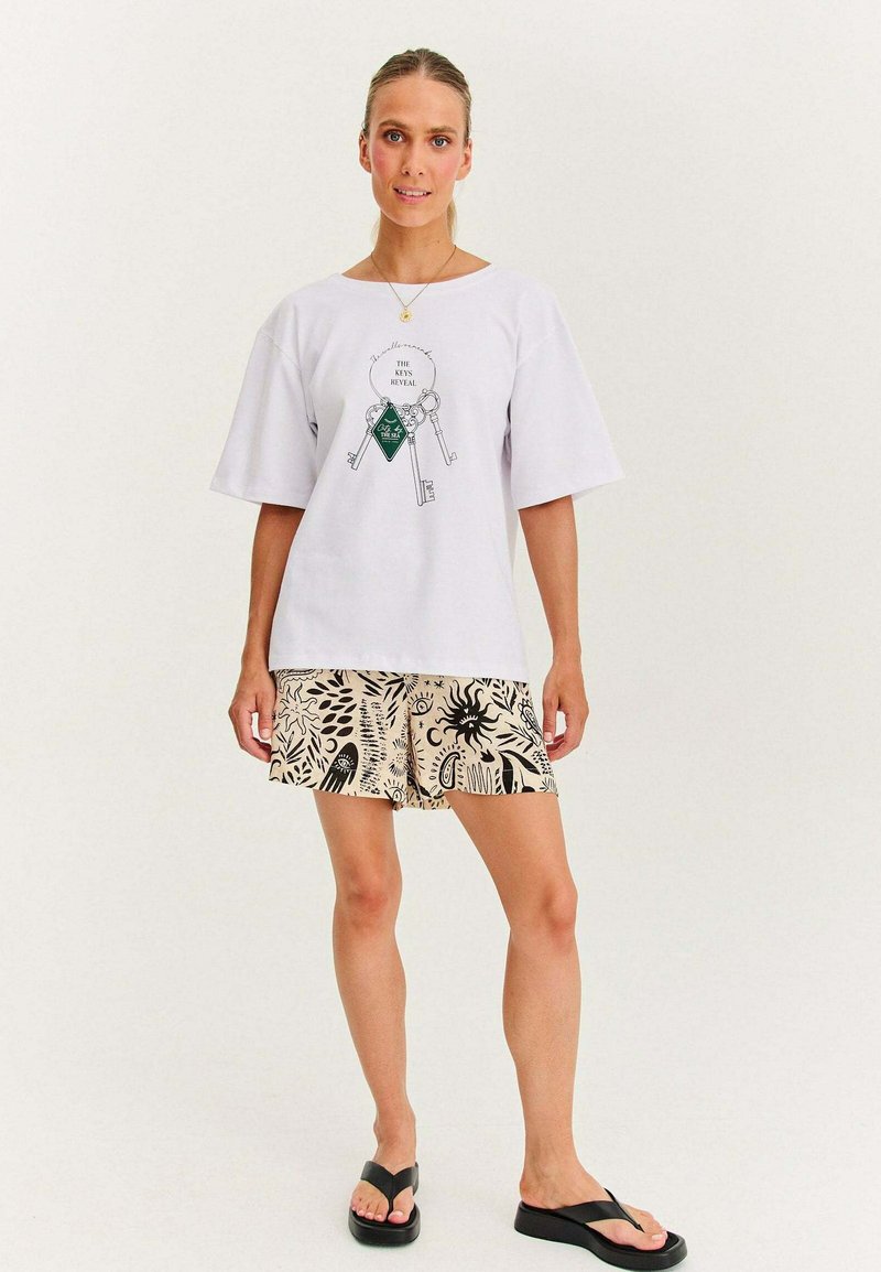 Kobieta ubrana w białą koszulkę z grafiką klucza i tekstem, beżowe szorty z czarnymi abstrakcyjnymi wzorami oraz czarne sandały na platformie, stojąca na białym tle.