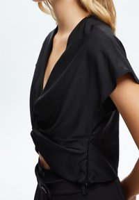 Černý top s krátkým rukávem a hlubokým výstřihem do V, s nařaseným pasem. Hladká látka s mírně lesklým povrchem. Nosit s přiléhavými černými kalhotami.