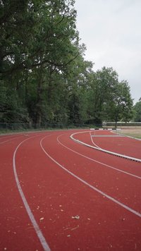 Piste d’athlétisme rouge qui tourne à droite avec des lignes blanches, entourée de grands arbres verts et un ciel nuageux en arrière-plan.