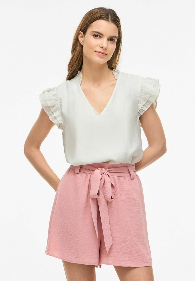 Femme portant un chemisier blanc à manches à volants rentré dans un short rose taille haute noué d'un nœud devant, debout les mains derrière le dos.