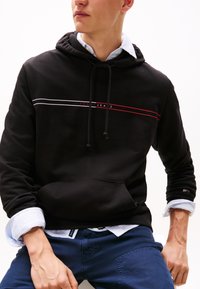Black hoodie with a drawstring, featuring white and red horizontal stripes and "TOMMY JEANS" logo. Cotton material with kangaroo pocket.