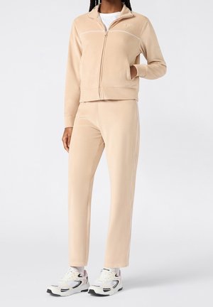 Persoon draagt een beige jack met rits en bijpassende broek met witte sneakers, staand tegen een effen lichte achtergrond.