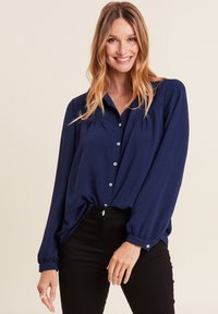 Blusa a maniche lunghe blu navy con chiusura a bottoni sul davanti, dettagli plissettati e polsini elasticizzati. Abbinata a pantaloni neri aderenti.