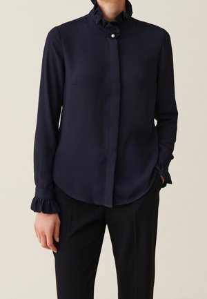 Vrouw die een marineblouse draagt met gerimpelde kraag en manchetten, parelknop bij de hals, en zwarte broek, staand met één hand in de zak.