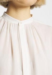 Gros plan d'une personne portant un chemisier blanc à col haut avec un seul bouton supérieur fermé.