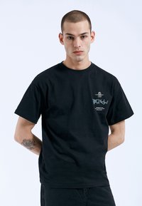 Černé bavlněné tričko s krátkým rukávem, s malým grafickým potiskem na hrudi. Jednoduchý design a hladká textura.