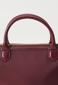 Burgundy handbag with smooth leather handles, hexagonal accents, and a gold zipper along the top. The body features a durable fabric texture.
