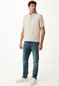 Light beige quilted vest with a high collar, featuring six buttons, two front pockets, worn over a short-sleeve shirt, paired with blue jeans and white sneakers.