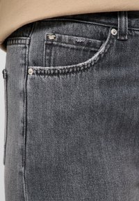 Gros plan sur la poche d'un jean en denim gris foncé avec des rivets argentés et un petit logo métallique en forme de cheval dans le coin supérieur.