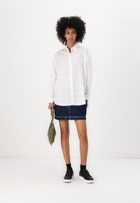 White long-sleeve shirt featuring large light gray "HUGO" text, paired with a dark denim skirt and black sneakers, holding a green bag.