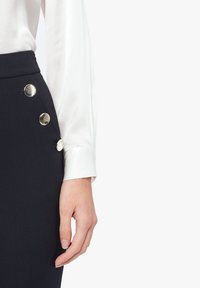 Jupe crayon marine avec des détails en boutons dorés sur le côté, associée à un blouse en satin blanc à manches longues. Texture lisse, design ajusté.