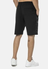 Schwarze Shorts mit einem elastischen Bund, seitlichen Taschen und einem strukturierten schwarzen Seitenpaneel mit einem Logo-Muster. Weiße Sneakers werden getragen.