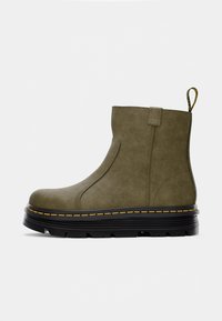 Bottine verte olive avec une tige en simili cuir lisse, des panneaux latéraux élastiques, des coutures jaunes et une semelle plate noire robuste.