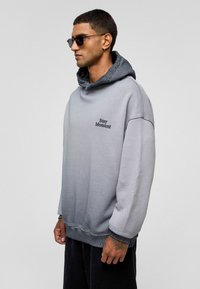 Grijze gradient hoodie met een gestructureerde afwerking, voorzien van een capuchon en "Stay Moment" borduring op de borst. Model draagt donkere broeken en een zonnebril.