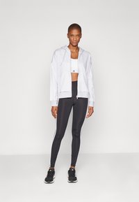 Sweatshirt cinzento com fecho, sobre uma t-shirt cropped branca, acompanhado de leggings pretas com riscas brancas laterais. Modelo calça sapatilhas pretas.