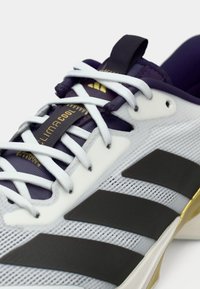 Buty sportowe z białą siateczkową cholewką, fioletowymi akcentami, złotym brandingiem i czarnymi gumowymi paskami. Posiadają białe sznurówki i wyściełany kołnierz.