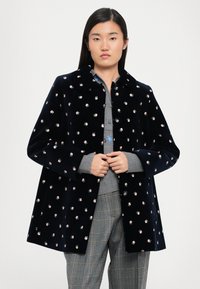 Merinovihreä samettitakki, jossa on hajallaan kukkakuvioita, kauluksella ja löysällä istuvuudella. Yhdistetty harmaan paidan ja ruututakki-housujen kanssa.