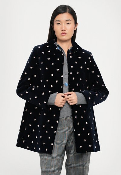 Granatowy aksamitny płaszcz z rozproszoną haftowaną florystyką, z kołnierzem i luźnym krojem. Połączony z szarym koszulą i w kratkę spodniami.