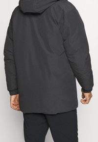 Persona che indossa un giubbotto invernale con cappuccio nero e pantaloni neri, vista da dietro con le mani rilassate ai lati.