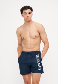 Marineblaue Badeshorts mit elastischem Bund, Kordelzug, Seitentaschen und weißem "Calvin Klein"-Logo am linken Bein.