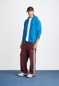 Sudadera azul con cremallera, camiseta blanca, joggers burdeos con franjas laterales, zapatillas grises y una gorra negra. Look de ropa deportiva casual.
