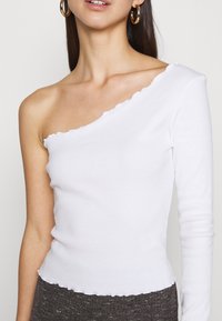 Vrouw die een witte, één-schouder lange mouw top met geschulpte randen en gouden creolen draagt, getoond van borst tot kin.