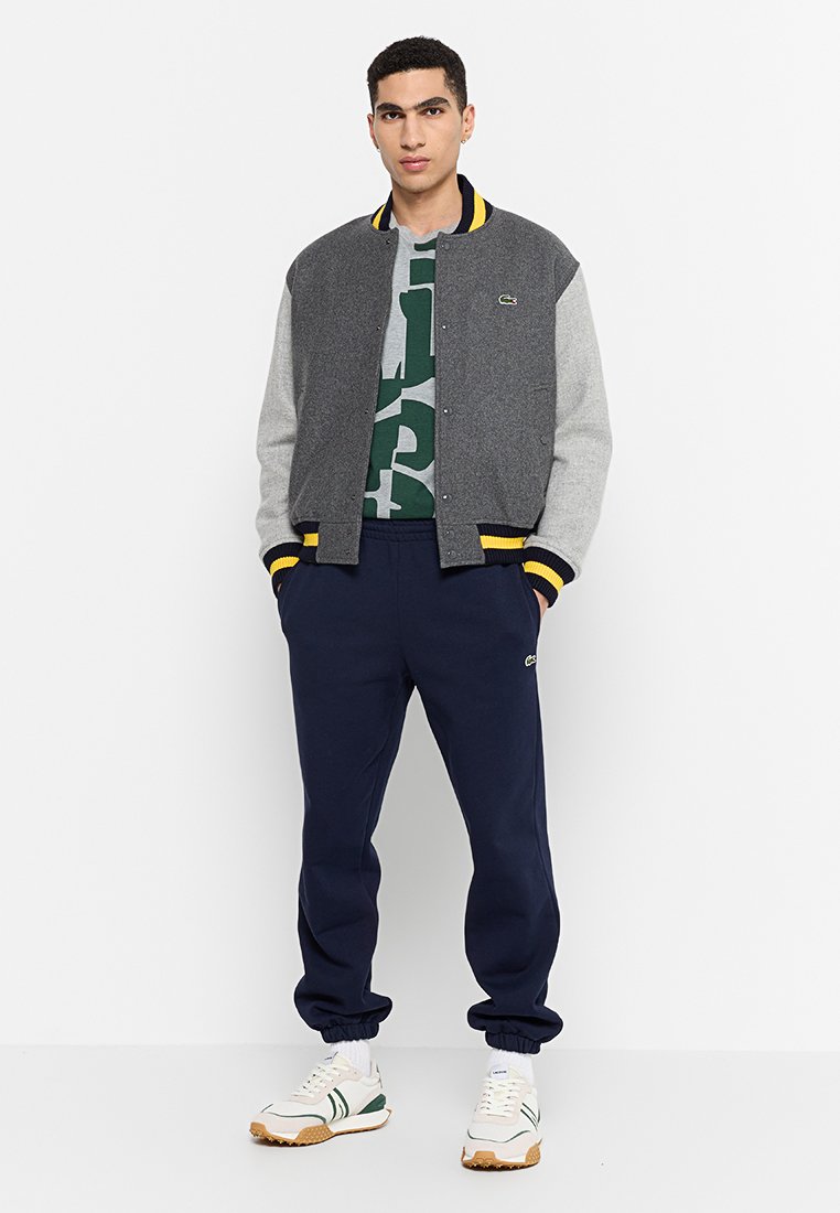 Jeune homme portant une veste universitaire grise, une chemise à motifs verts et gris, un pantalon de jogging bleu marine et des baskets blanches, se tenant les mains dans les poches.