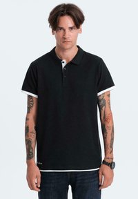 Černé polo tričko s bílým límcem a manžetami, vyrobené z bavlny. Má zapínání na tři knoflíky a je kombinováno s tmavými džíny.