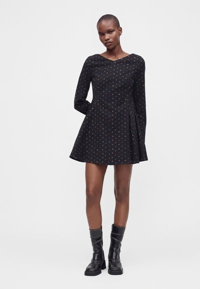 Femme portant une robe noire à manches longues avec de petits imprimés colorés et des bottes noires mi-mollet, debout devant un fond clair uni.