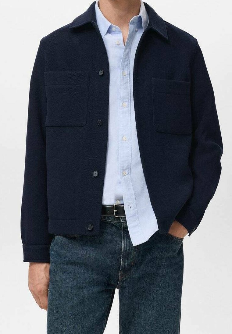 Homme portant une veste bleu marine foncée boutonnée avec deux poches poitrine sur une chemise bleu clair et un jean bleu foncé, main dans la poche, de la taille vers le haut.